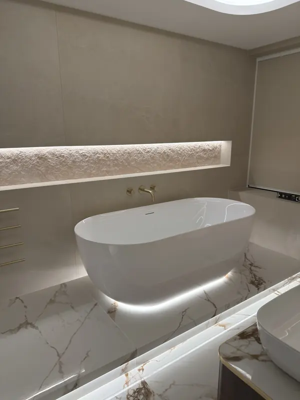 Freestanding bath on a marble-effect plinth lit along its edge, beneath a long lit stone niche