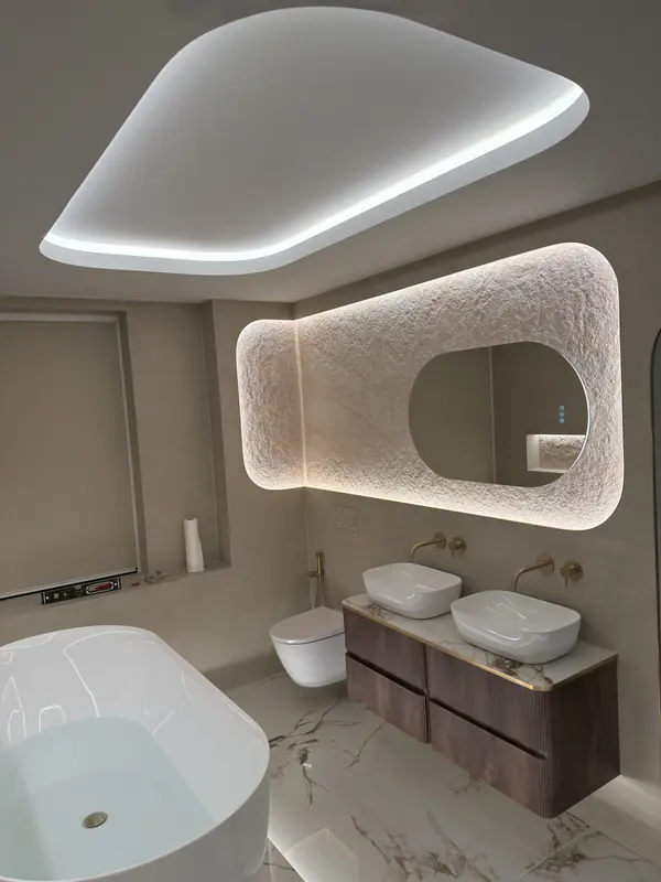 Lit curved ceiling coffer above a backlit textured stone panel with an inset mirror and a walnut double vanity