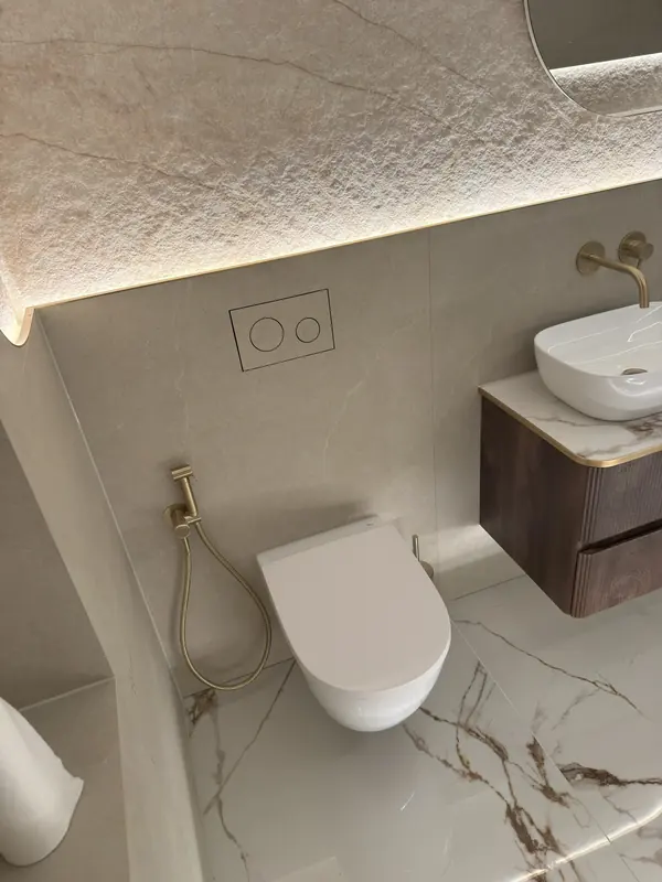 Wall-hung WC with a brass douche spray beside a walnut vanity, under a backlit stone panel